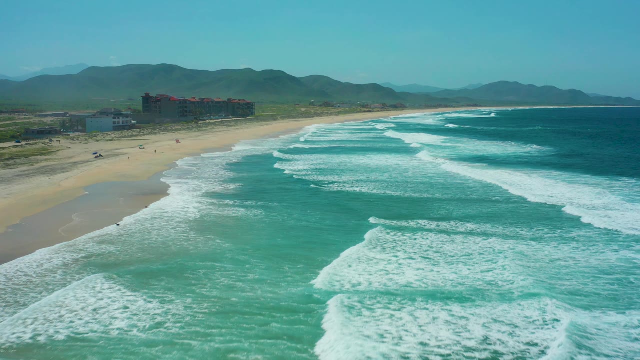 At Cerritos Beach, foamy surf fills more of the foreground while the sandy beach curves left toward low buildings and hazy green mountains under a clear sky, with small people visible.