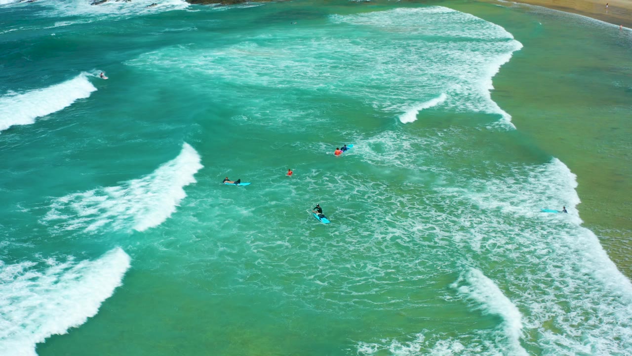Aerial frame of Cerritos Beach surf with small visible surfers, blue boards, and a wide sheet of whitewater moving across green-blue ocean water.