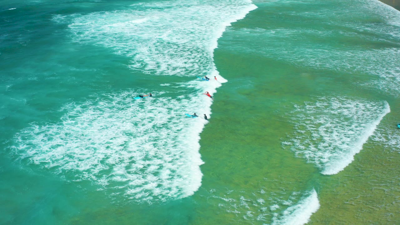 Overhead aerial frame at Cerritos Beach, Baja California Sur, showing turquoise ocean, diagonal white surf, green shallows, and several visible surfers on boards.