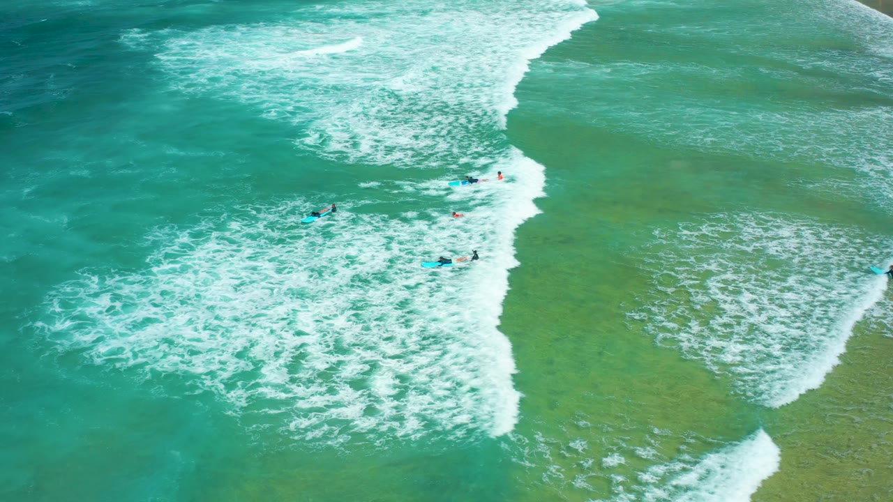 Aerial view at Cerritos Beach with several visible surfers separated across turquoise water while foamy white surf expands across the center of frame.