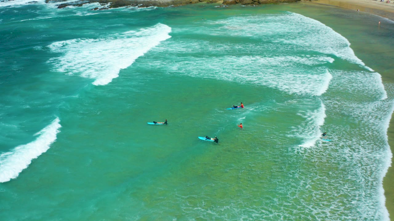 Aerial view at Cerritos Beach, Baja California Sur, with several surfers on blue boards spaced across shallow turquoise water as white surf curves in from the upper frame.