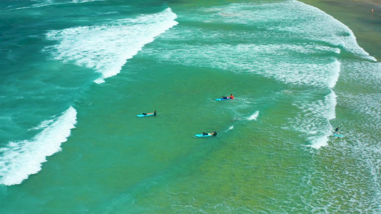 Aerial Cerritos Beach view with three main surfers visible below sweeping whitewater, including one darker figure on a blue board and another red-clad figure near the center.
