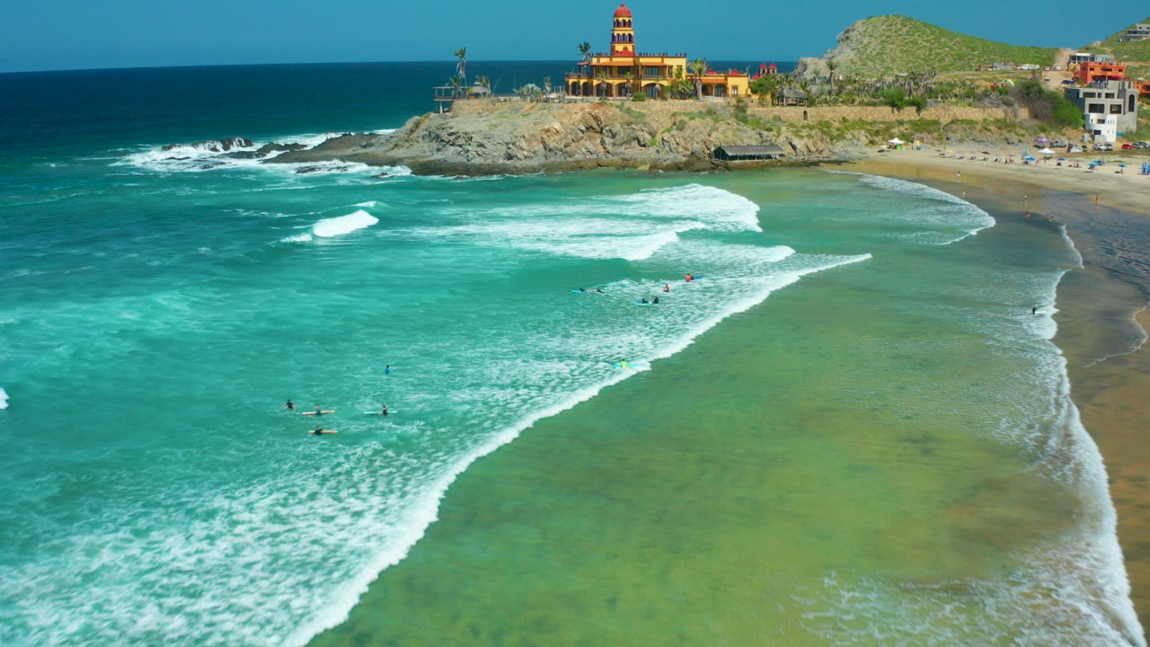 Aerial frame from Cerritos Beach, Baja California Sur, showing turquoise water, white breaking waves, small people in the surf, a sandy beach, and buildings on a rocky headland.