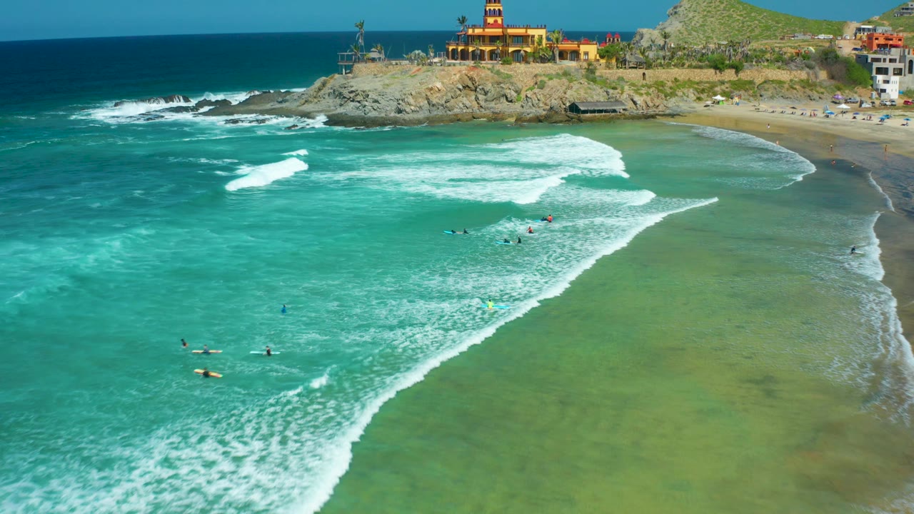 Aerial frame at Cerritos Beach showing larger white wave patches across turquoise water, tiny people with surfboards near the center, sand along the right edge, and buildings above the rocks.