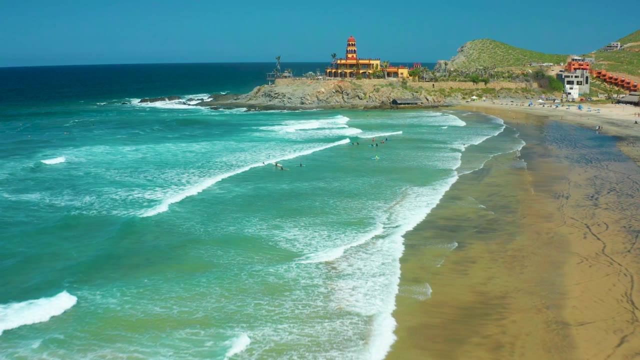 Cerritos Beach aerial view with long white foam bands crossing green-blue water, small swimmers near the center, and the lighthouse structure set back on the rocky point.