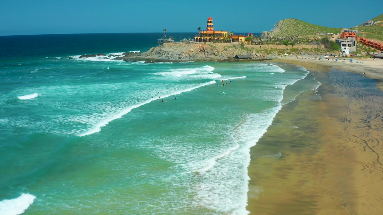 At Cerritos Beach, a broad white wave line curves toward the sand while scattered swimmers remain small in the water and the lighthouse stands above the rocky shoreline.