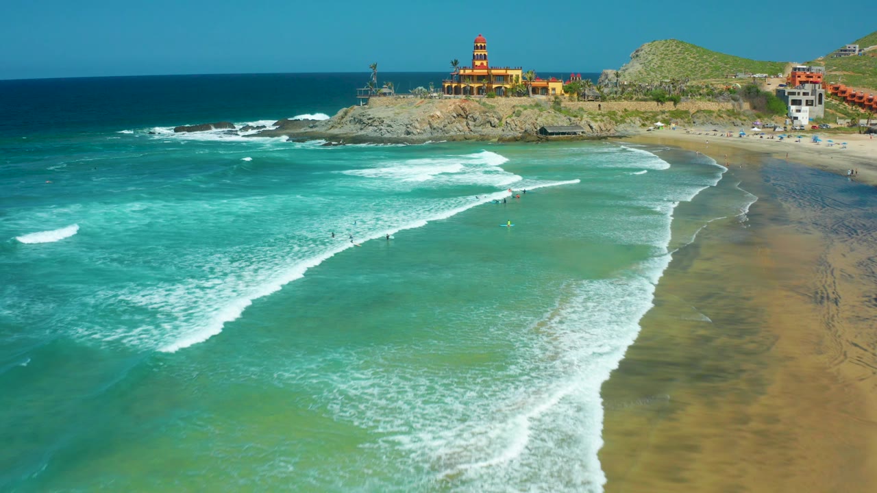 Turquoise water and foamy surf beside a sandy shore with a lighthouse in back.