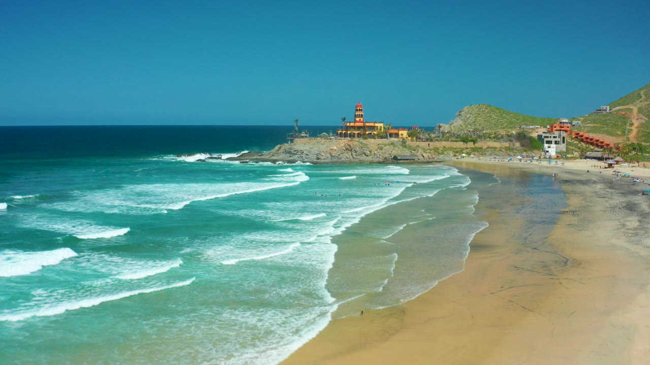 Wide aerial frame from Cerritos Beach, Baja California Sur, showing turquoise surf lines crossing wet sand with the lighthouse point and hillside in the distance.