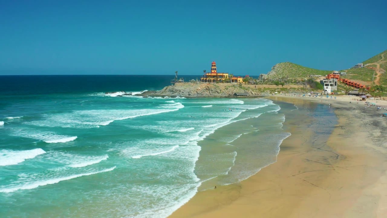 Aerial beach view with white wave bands, wet sand, a rocky point, and lighthouse under clear sky.