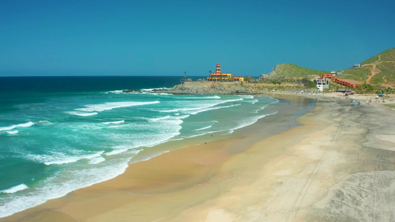 Wide aerial beach view with white wave foam, sand, rocky shoreline, and lighthouse.