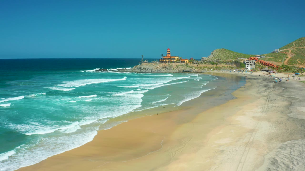 At 0:02, the surf spreads farther across the sand at Cerritos Beach, with the lighthouse, rocky point, beach buildings, and hillside still visible under clear light.