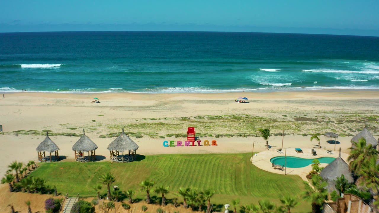 Opening aerial frame at Cerritos Beach, Baja California Sur, showing thatched huts, green lawn, a small pool, colorful CERRITOS lettering on sand, turquoise ocean, and no visible people.