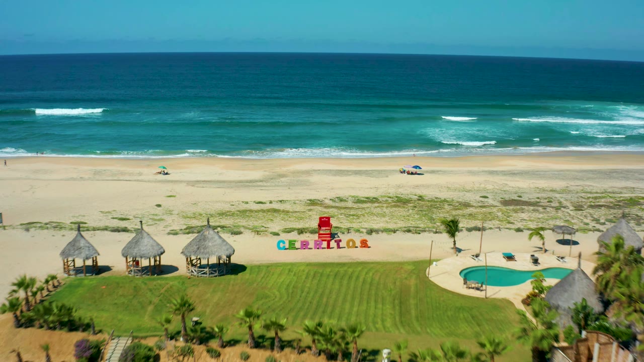 Aerial beach view with thatched huts, pool, lawn, sand, and surf.