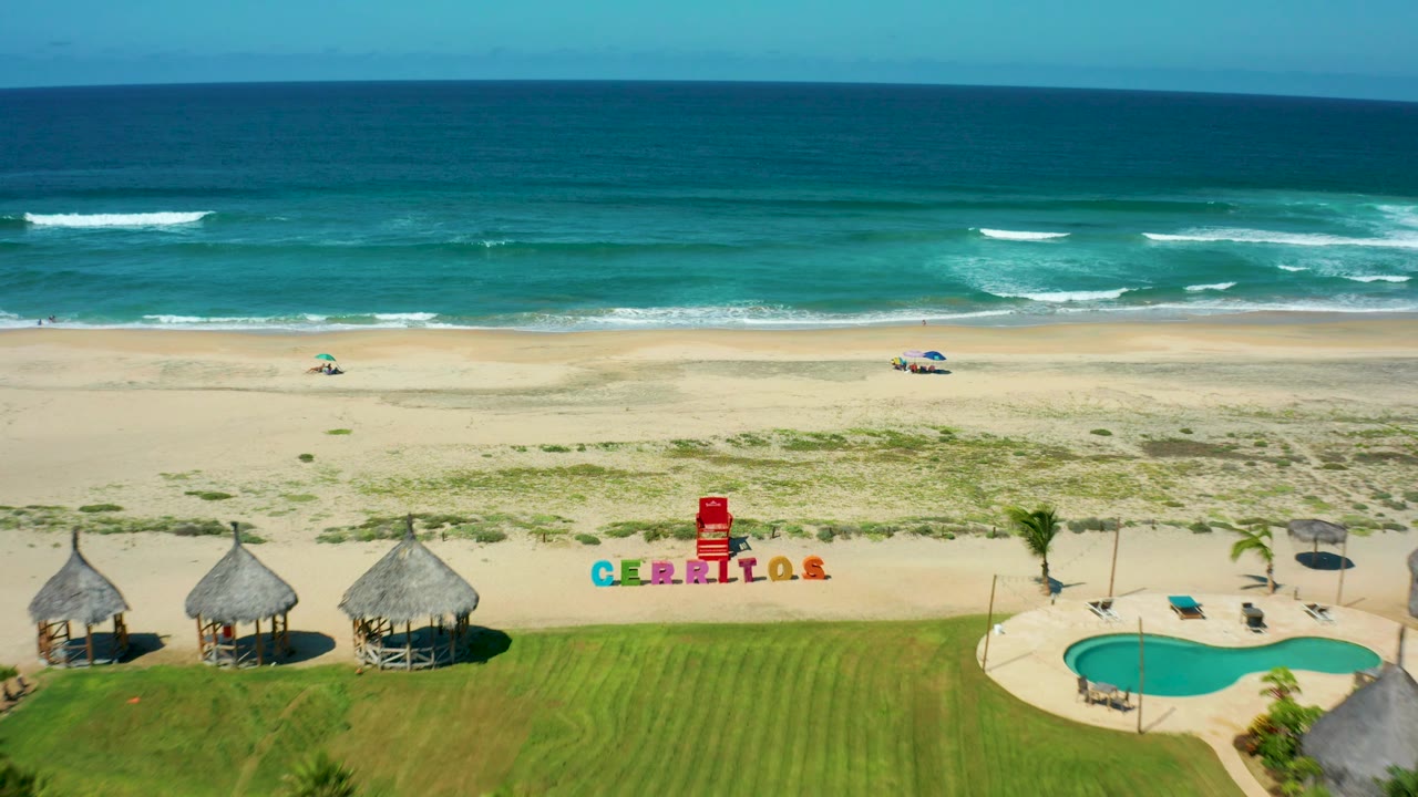 High aerial view over Cerritos Beach with thatched shade huts, a lawn area, a pool at frame right, pale sand, rolling surf, and no visible people.