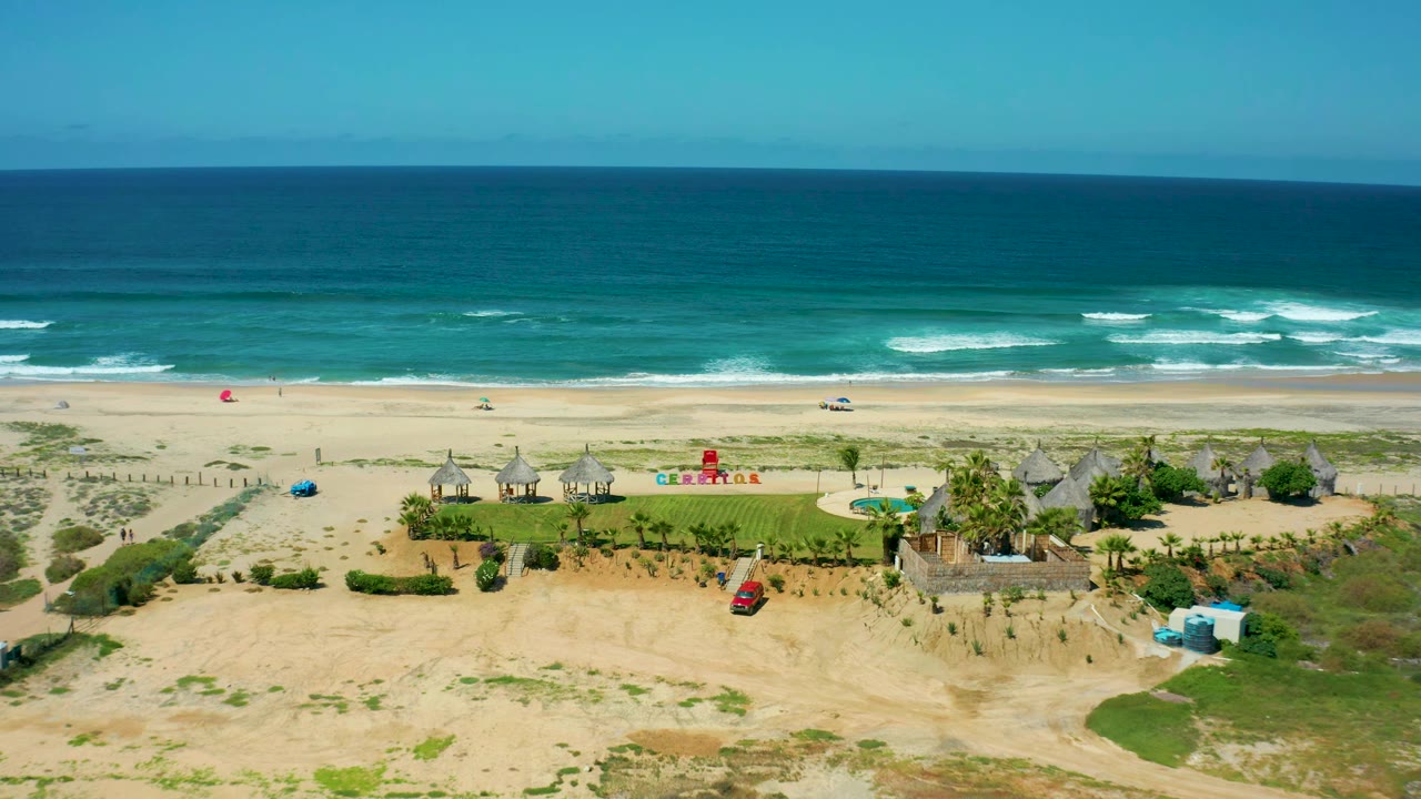 Aerial view at Cerritos Beach with the red car lower in frame, green resort lawn and thatched huts in the middle, breaking waves behind the beach, and no visible people.