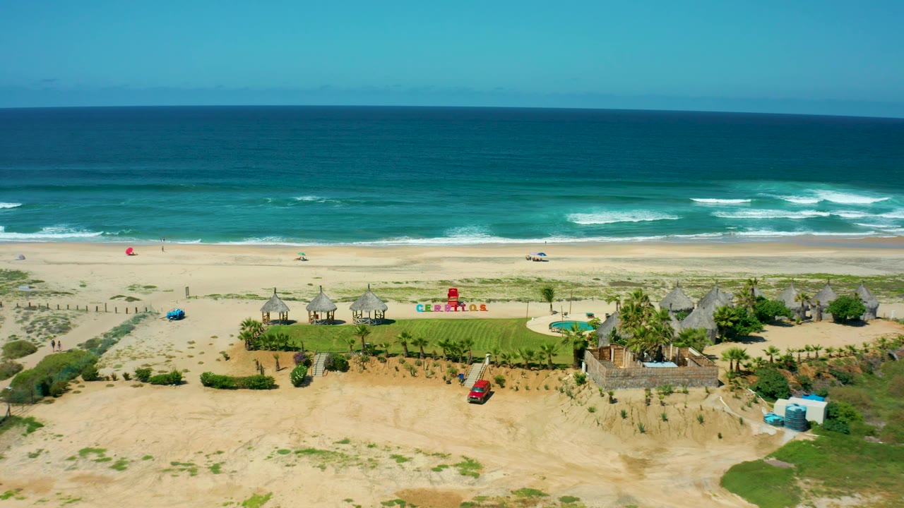 Cerritos Beach aerial frame showing a broad ocean horizon, white surf, sandy beach, resort cabanas, a pool beside green lawn, a red car near the lower edge, and no visible people.