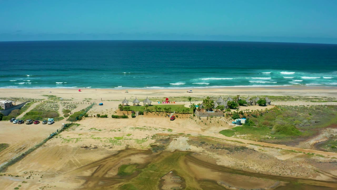 Wide aerial view of beach huts, vehicles, sand, and ocean.