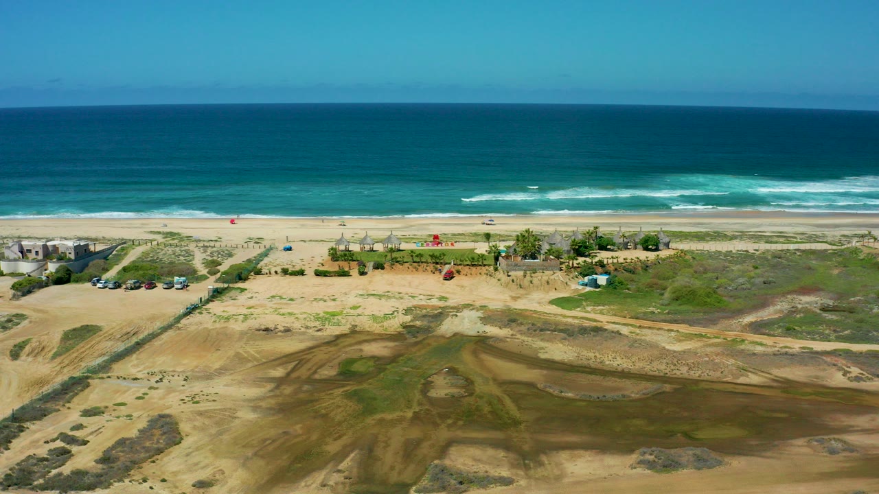 Opening aerial frame over Cerritos Beach, Baja California Sur, showing blue ocean, white surf, sandy beach, dune ground, scattered buildings, parked vehicles, and no visible people.