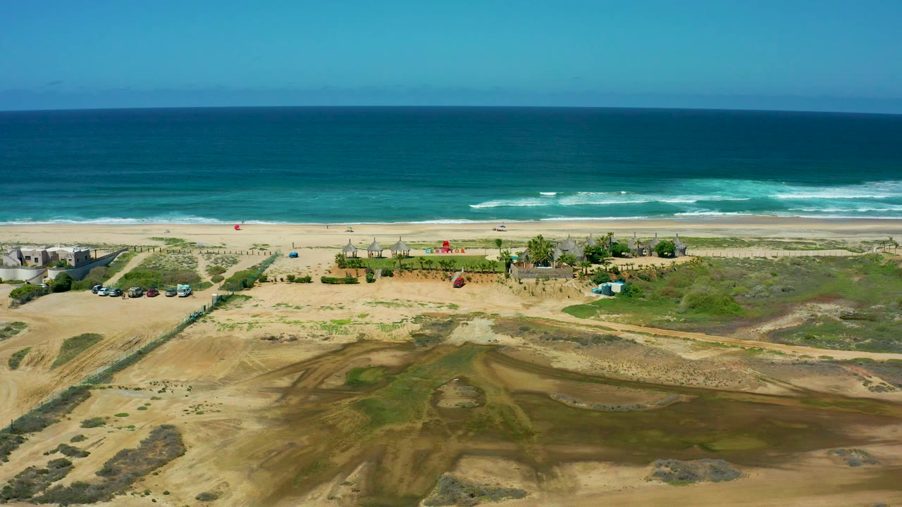 Cerritos Beach aerial view with a broad band of blue water, white foam along the shore, pale sand, scrubby dunes, scattered buildings, parked vehicles, and no visible people.