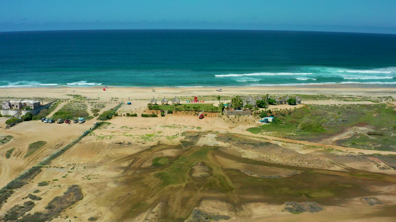 Aerial frame over Cerritos Beach, Baja California Sur, with tan dunes and green patches below, small beach structures near the sand, blue ocean beyond, and no visible people.