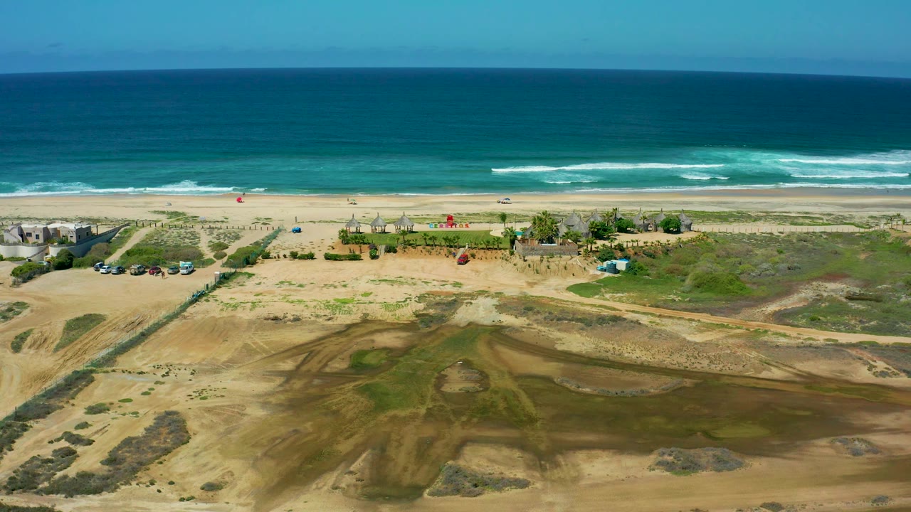 A wide Cerritos Beach frame shows dune ridges and scrub in the foreground, a narrow beach zone with small structures, rolling blue surf behind it, and no visible people.