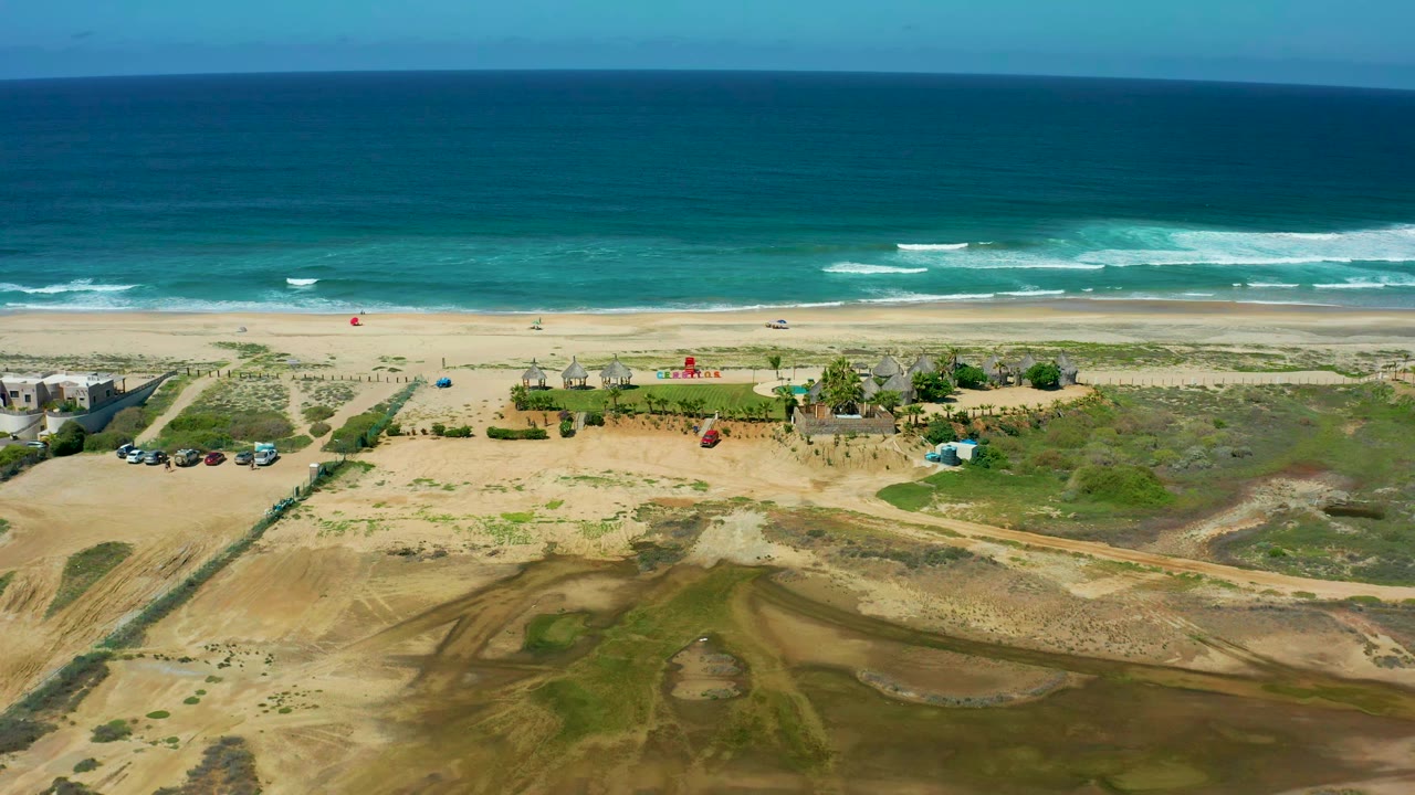 Second frame of Cerritos Beach shows teal water and breaking waves behind a sandy beach, with vehicles clustered near a dirt access path and no visible people.