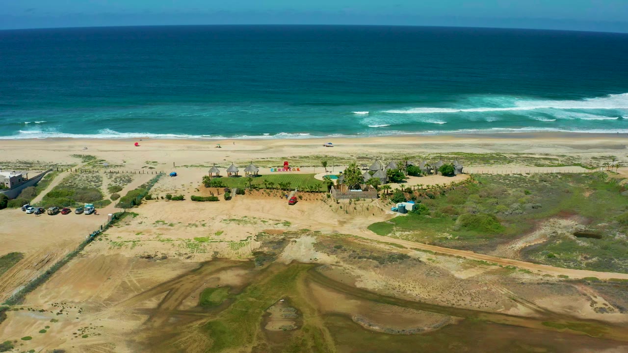 Aerial frame over Cerritos Beach, Baja California Sur, with blue ocean, white surf, sandy beach, dune vegetation, parked vehicles, shore structures, a small red kite near the sand, and no visible people.