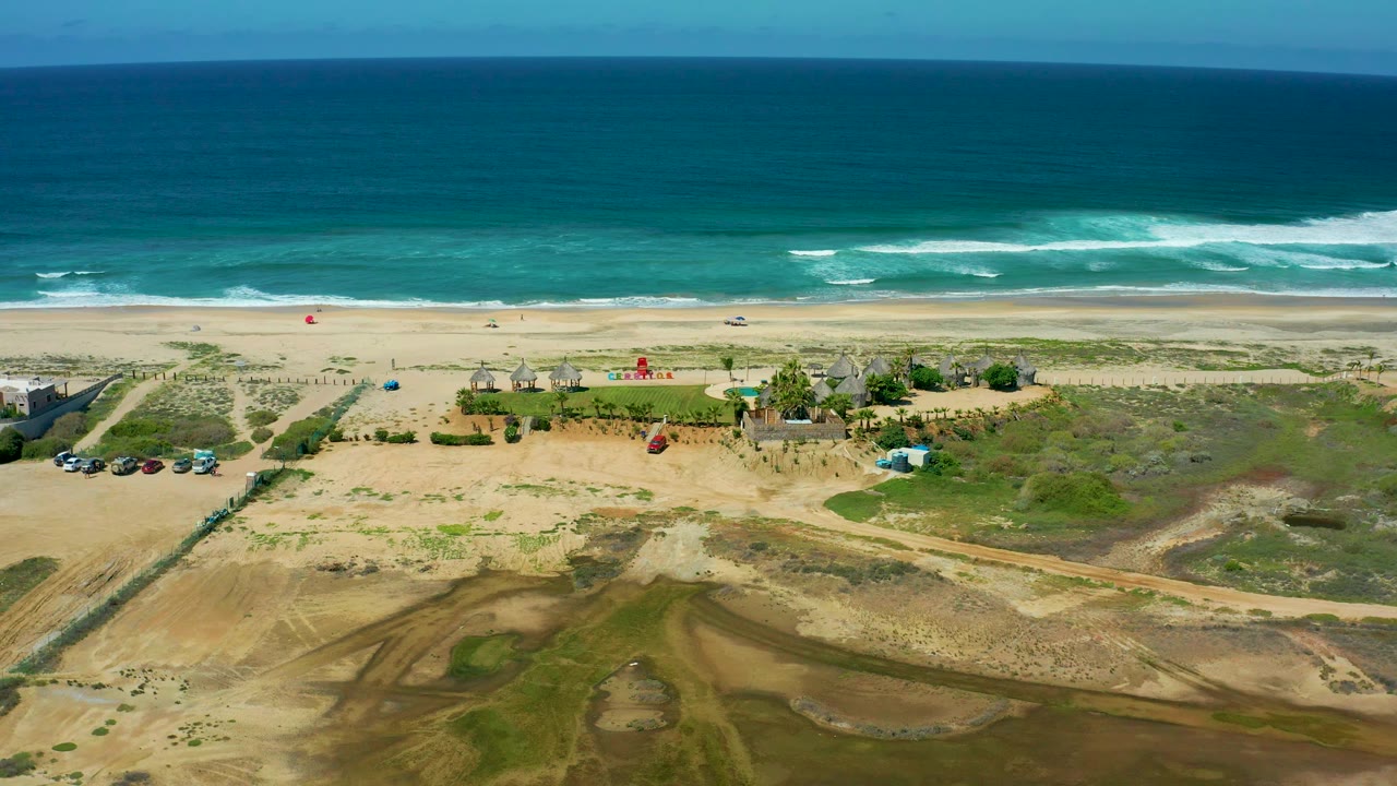 Cerritos Beach aerial frame with the ocean horizon, foamy wave lines, open sand, low dune vegetation, beach access tracks, parked vehicles, small structures, and no visible people.