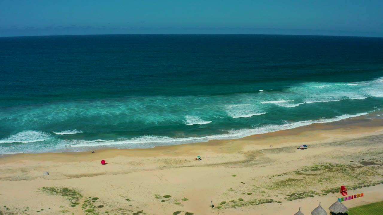 Aerial frame over Cerritos Beach in Baja California Sur showing open sand, blue-green surf bands, small huts near grass, red shore flags, and no visible people.