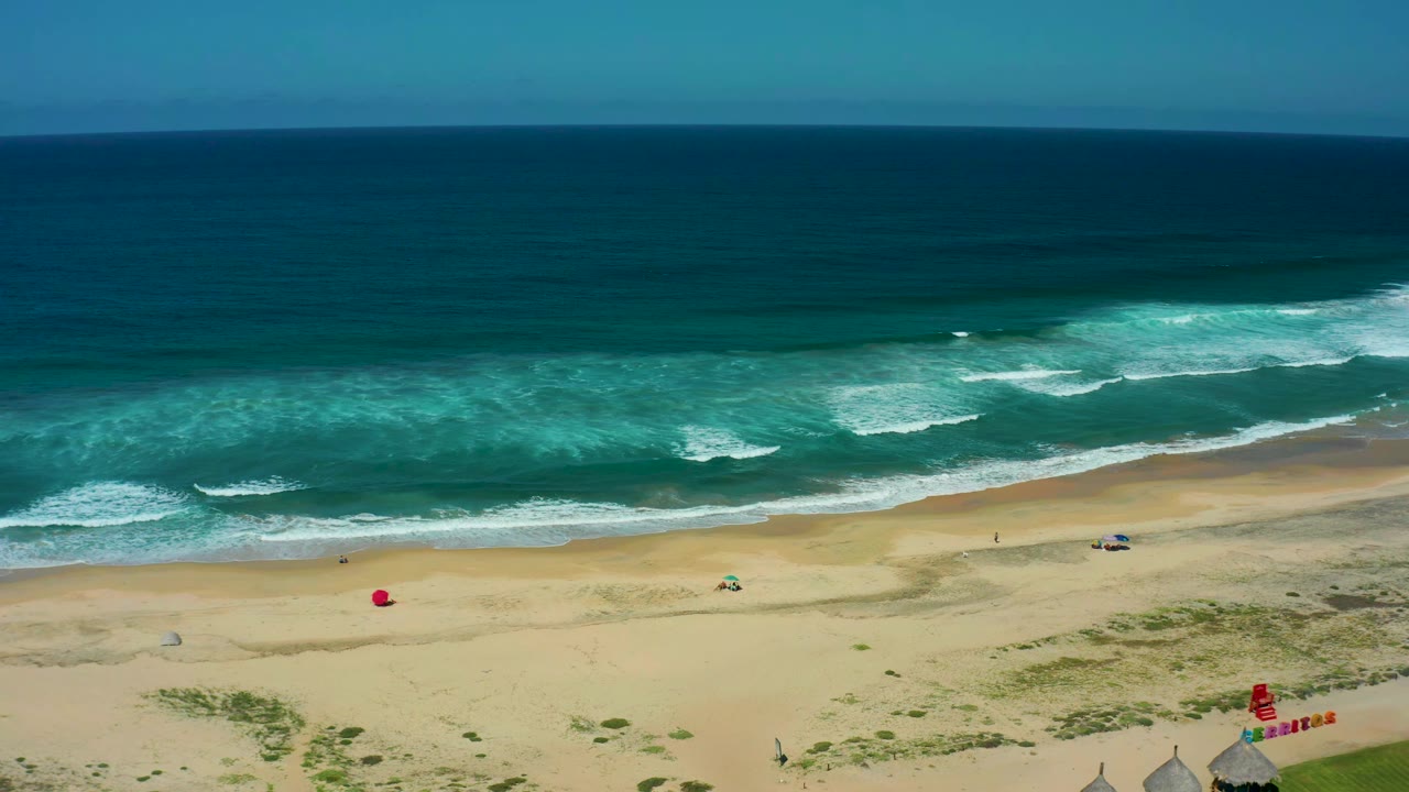 Wide aerial beach view with rolling surf, red flag, blue object, huts, and grass.