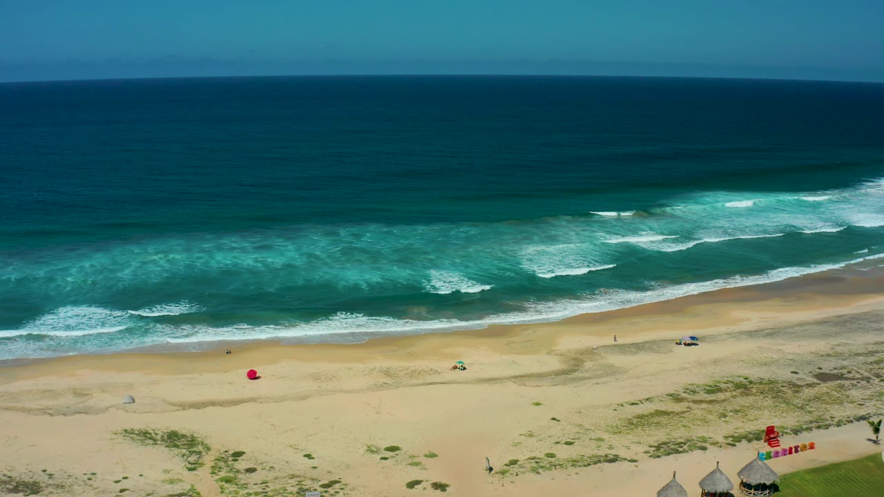 Aerial view of Cerritos Beach with stronger white wave breaks near the right side, open tan sand below, red flags near the huts, and no visible people.