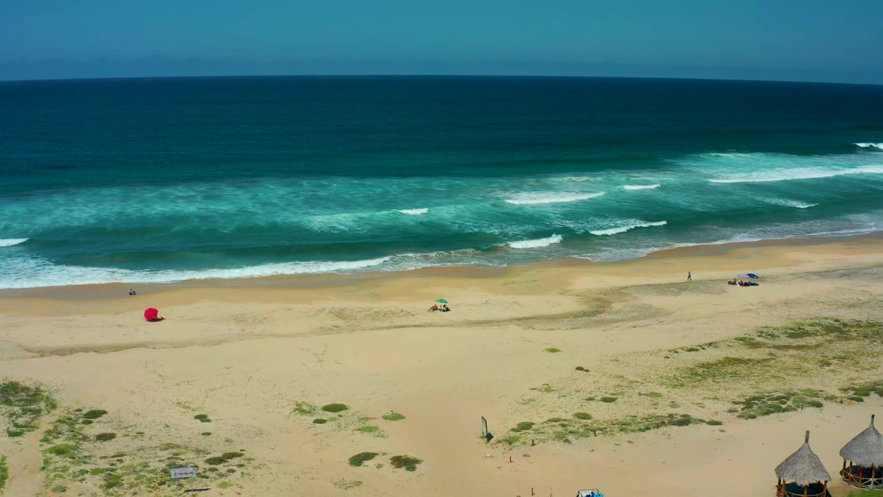 Wide aerial frame of Cerritos Beach with dark blue ocean, turquoise surf, tan sand, a red umbrella or marker, a small green umbrella, and tiny beachgoers visible.