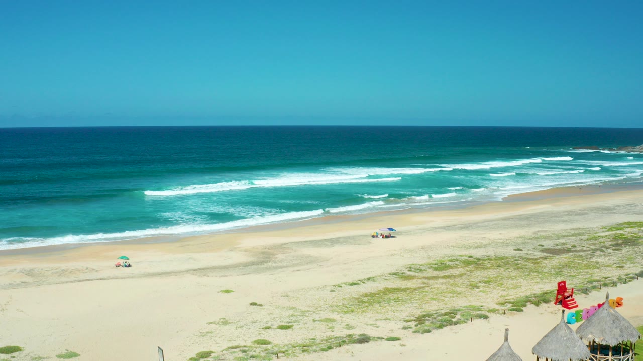 Cerritos Beach in Baja California Sur seen from above, with turquoise waves, broad sand, small umbrellas, and thatched huts partly visible at the bottom edge.