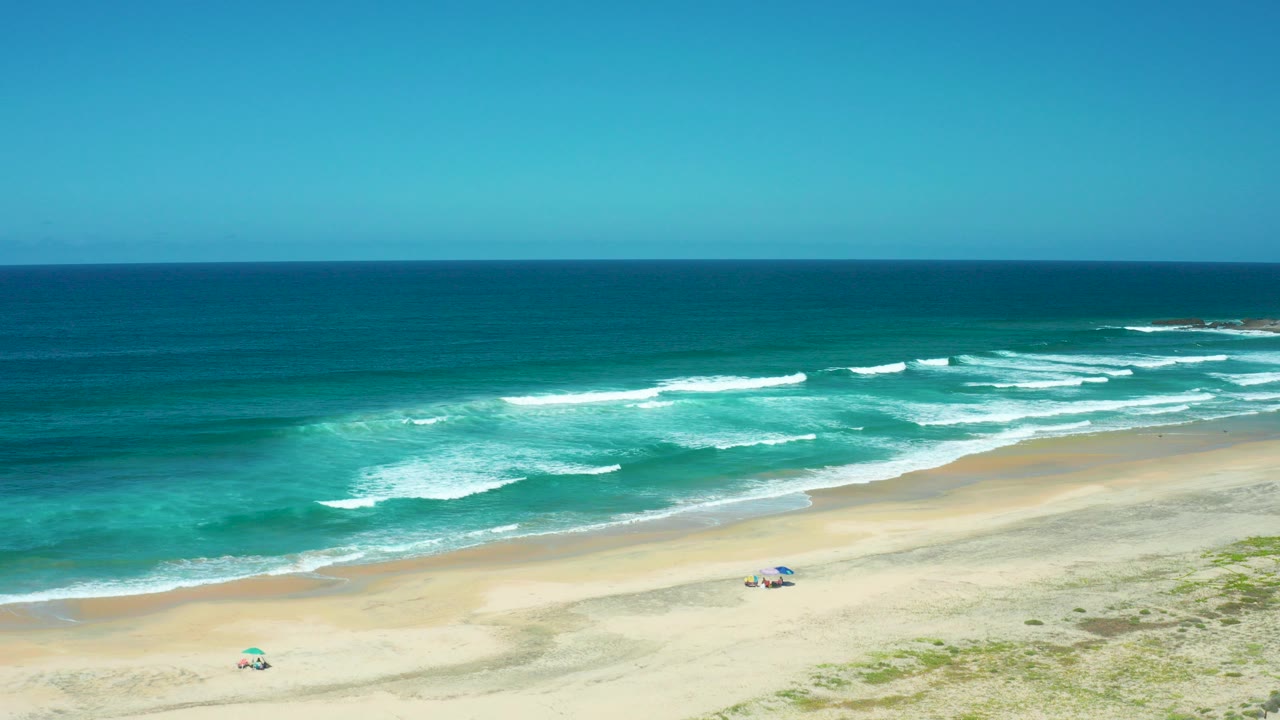 Cerritos Beach, Baja California Sur, appears from above with turquoise water, white wave lines, pale sand, two small umbrellas, and tiny beachgoers near the shoreline.