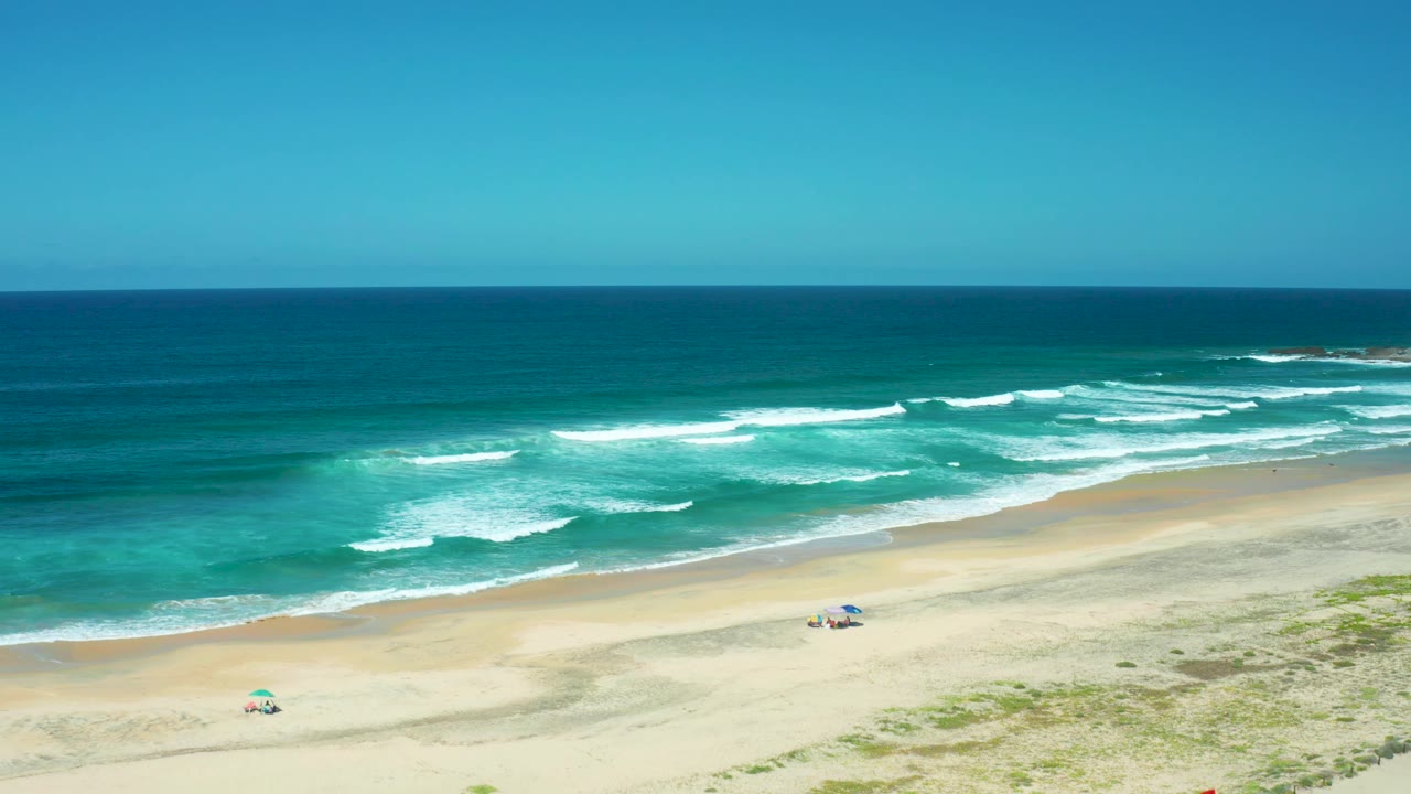 Cerritos Beach is shown with broad open sand, turquoise ocean, long white wave breaks, a blue umbrella on the mid-beach, and a small umbrella lower left.