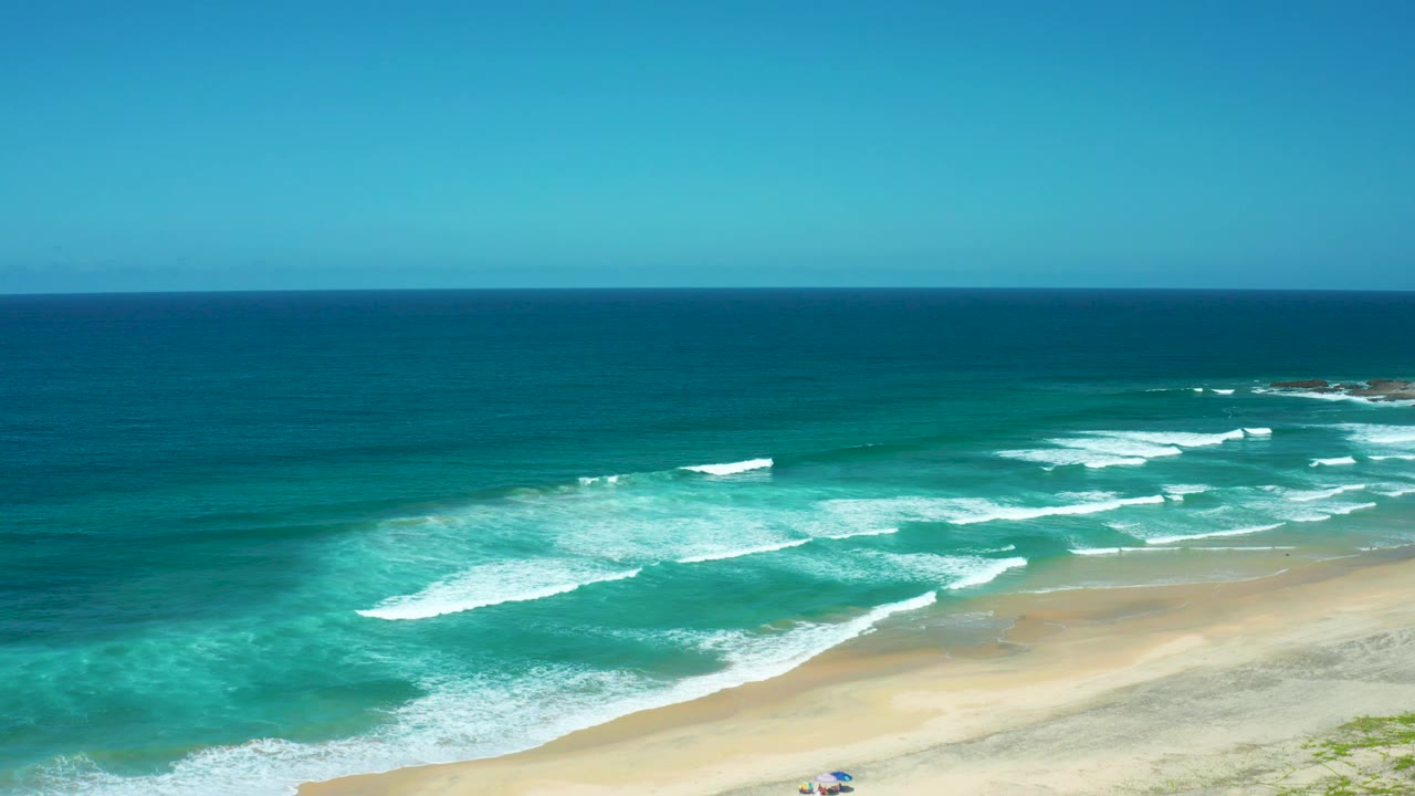 Elevated view over Cerritos Beach with bright turquoise shallows, foamy breaking waves, a wide sand strip, and a small umbrella shape near the lower beach.