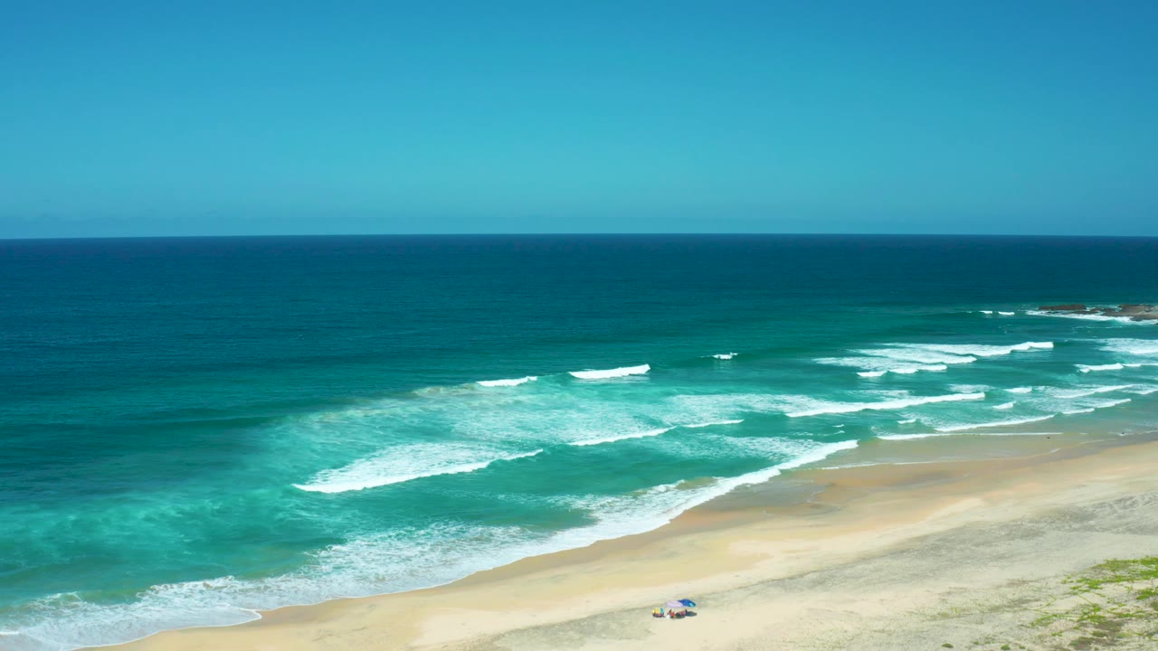 Aerial coastal view with surf, sand, and a small beach umbrella.