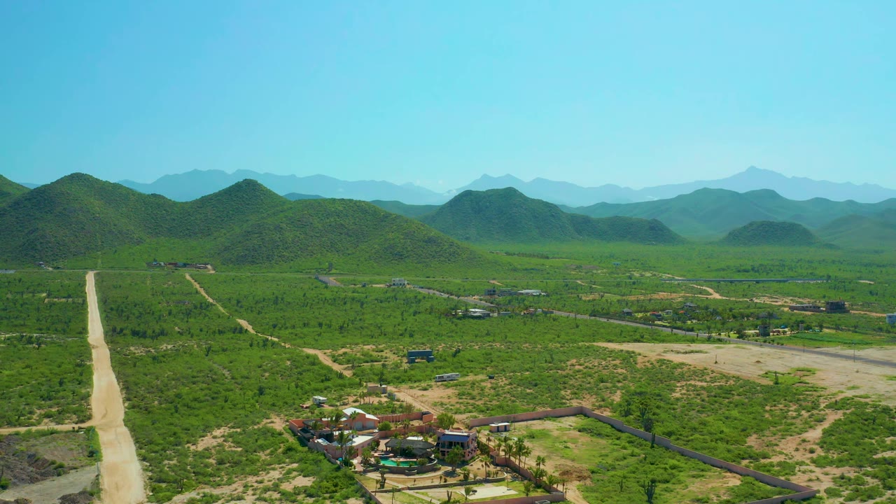 Aerial valley view with dirt road, small buildings, green hills, and blue sky.