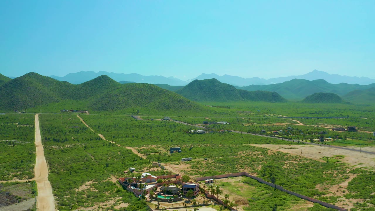 Aerial view in Cerritos Beach, Baja California Sur with a pale road at left, tan tracks through green fields, small structures in the lower foreground, and no visible people.