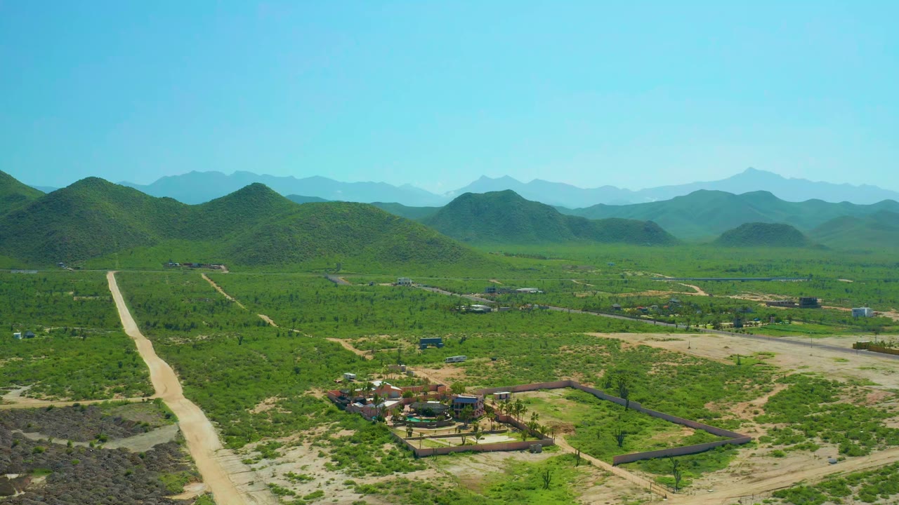 Aerial view in Cerritos Beach, Baja California Sur with the dirt road running up the left side, a small building cluster near the lower center, green scrubland, hazy hills, and no visible people.