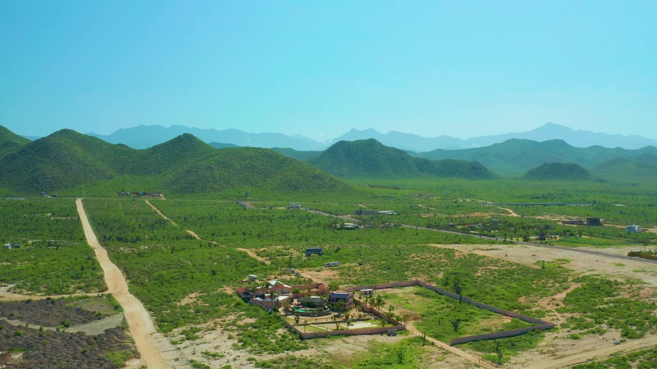 Wide drone frame over Cerritos Beach countryside showing the dirt road at left, low buildings lower in frame, patchy green vegetation, layered mountain ridges, and no visible people.