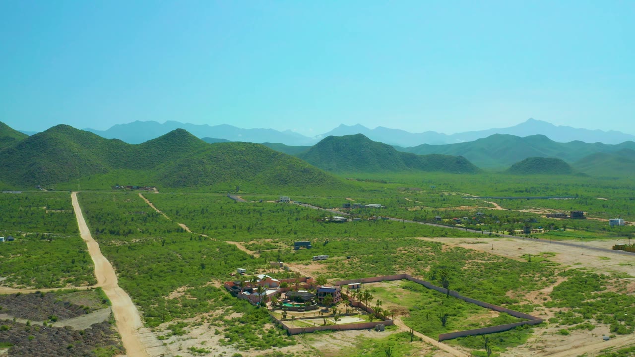 Cerritos Beach, Baja California Sur aerial frame with the straight dirt road along the left edge, scattered structures below, broad green terrain, soft blue hills, and no visible people.