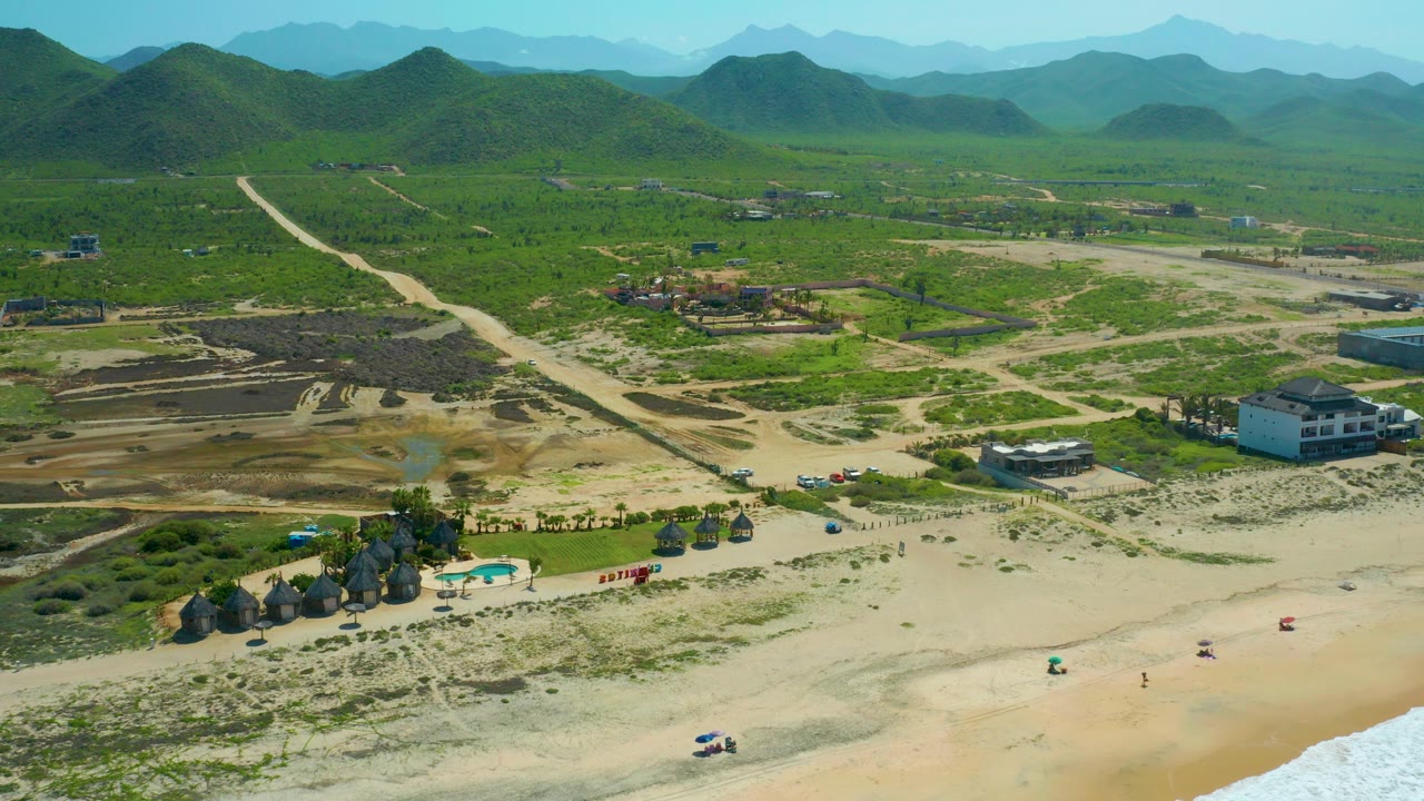 Aerial frame from Cerritos Beach, Baja California Sur, showing pale sand in front, thatch huts and a small pool below, dirt roads and low buildings beyond, and green hills across the horizon.