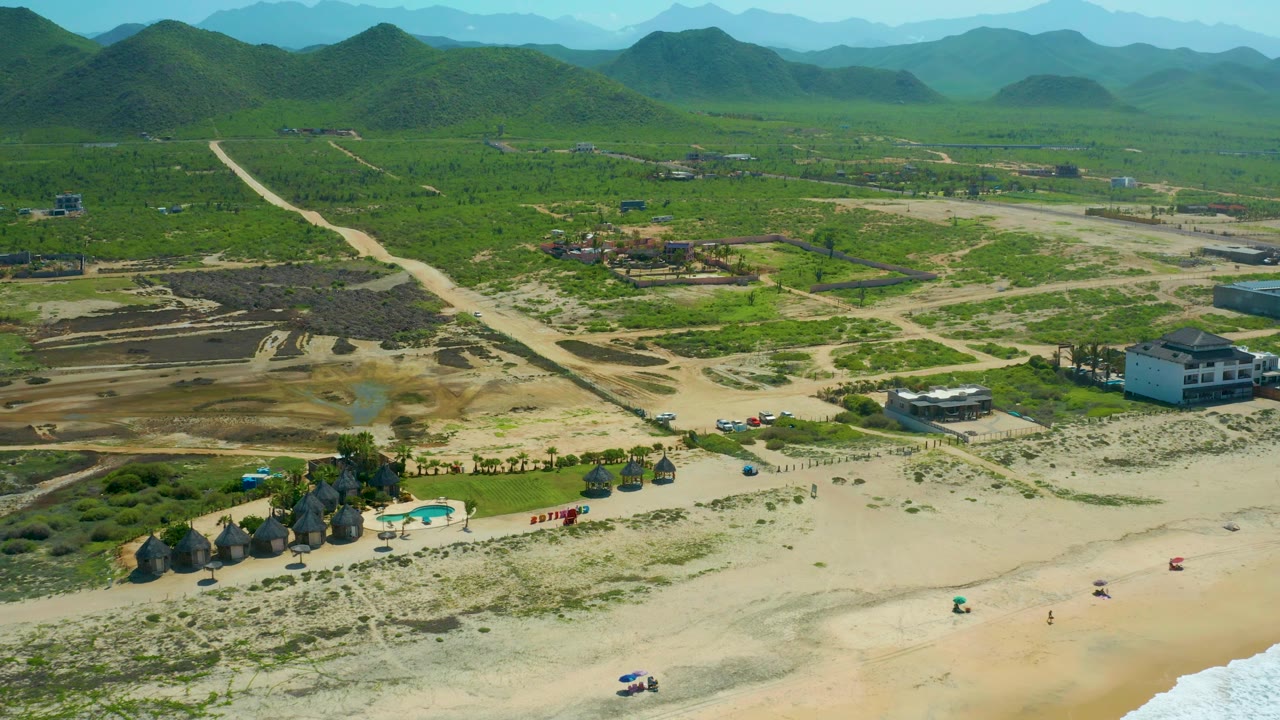 Aerial frame over Cerritos Beach with beach sand and a small surf line at the bottom, huts and pool near the lower left, dirt lots and roads in the center, and layered green hills beyond.