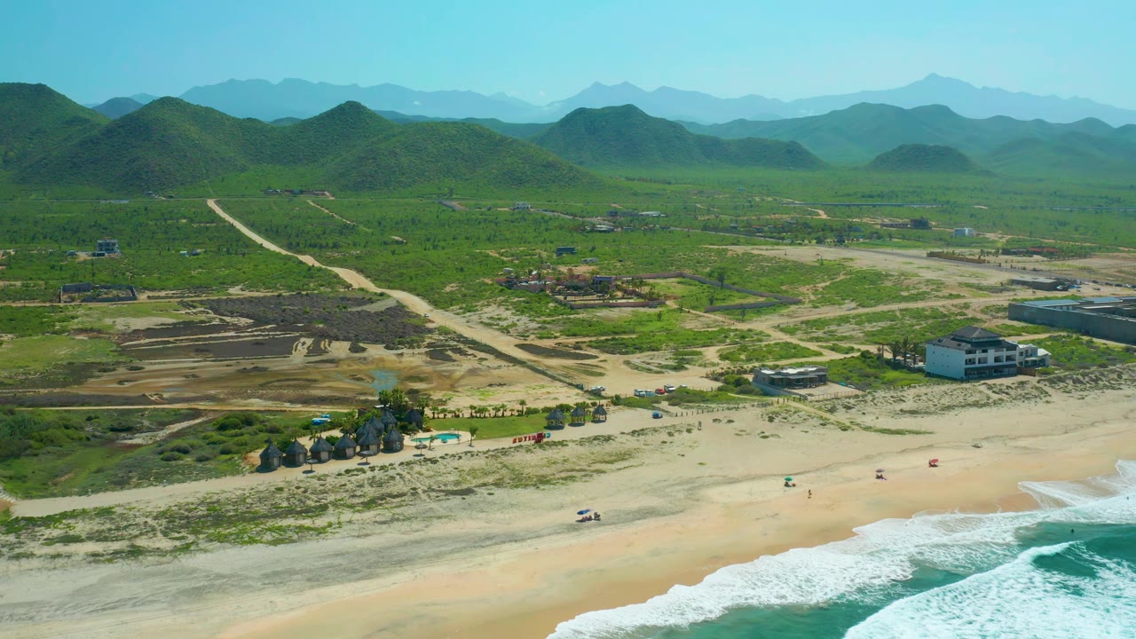 Wide drone view of beach road, surf line, huts, buildings, and hills.