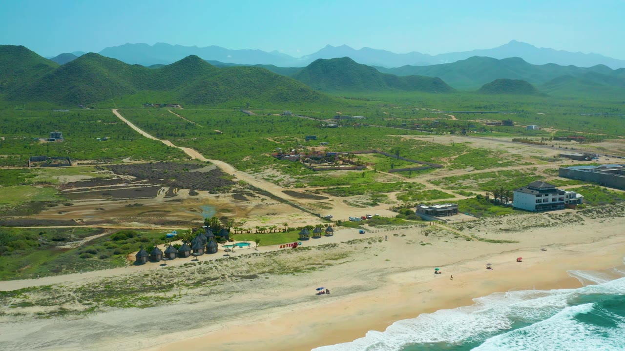 Aerial view over Cerritos Beach with waves entering at the lower right, a pale dirt road running inland, scattered people on the sand, low buildings, green hills, and hazy mountain layers.