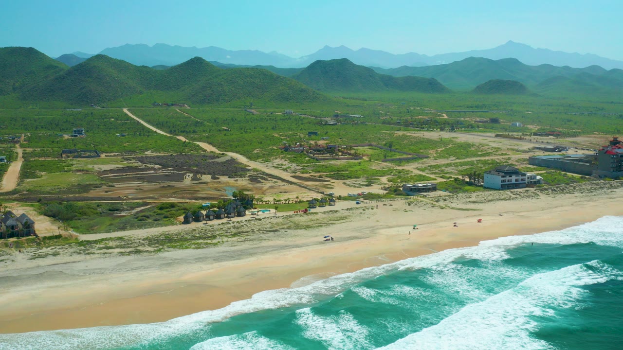 High aerial view over Cerritos Beach in Baja California Sur with turquoise waves in the foreground, pale sand, tiny beach activity, scattered buildings, dirt paths, green hills, and distant blue mountains.