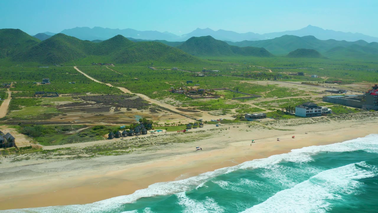 Cerritos Beach aerial view with foamy turquoise water crossing the lower right, sandy beach activity at small scale, a cluster of shore buildings, dirt paths, green hills, and layered mountains in the distance.