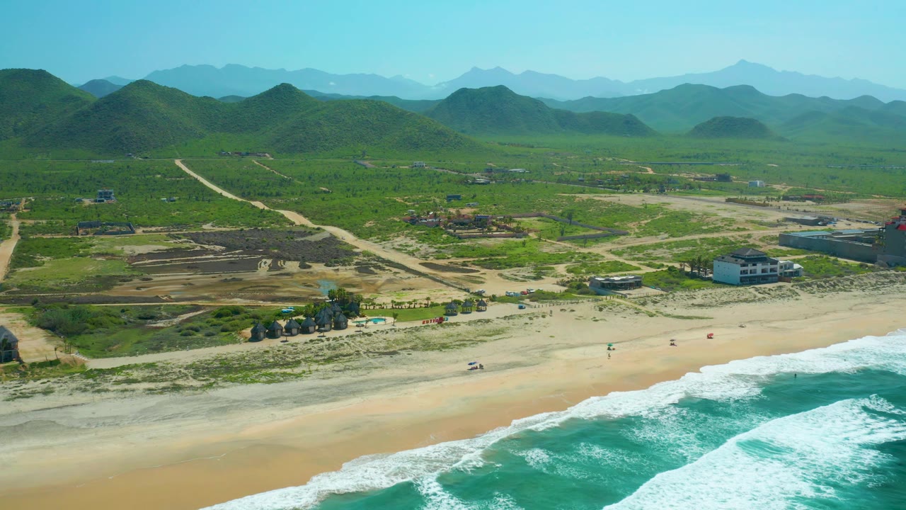 Drone view of beach waves, pale sand, small shore structures, dirt lots, and green mountains.
