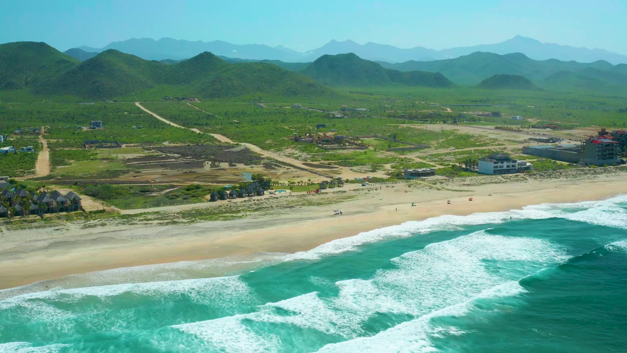 Cerritos Beach aerial frame with white surf advancing across turquoise water, a pale sand strip, scattered buildings, inland dirt roads, green hills, and no visible people.